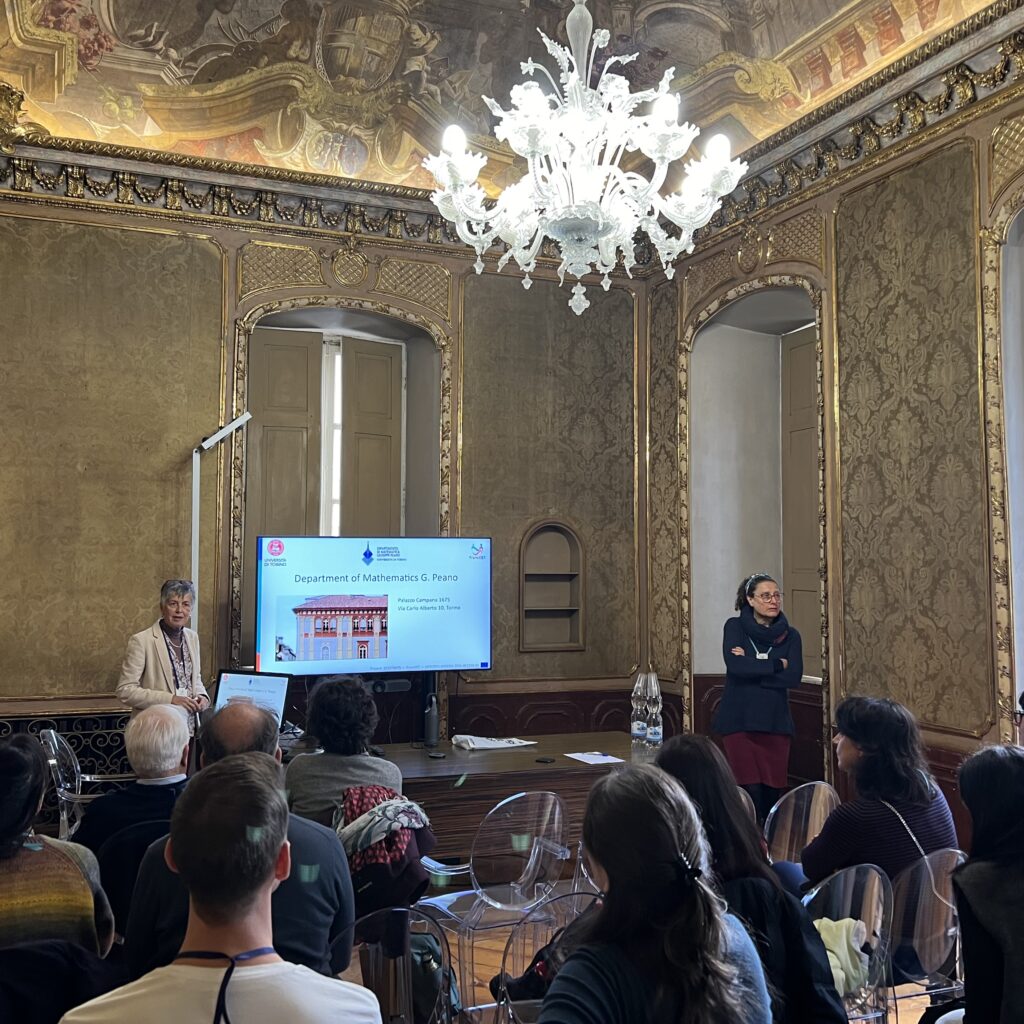 Two women standing presenting the University of Turin Department of Mathematics to an audience in an elegant baroque room.