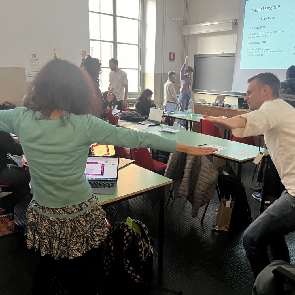A classroom filled with workshop participants performing dance moves in front of laptops.