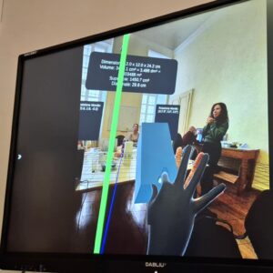 A television screen showing the view of an office through an augmented reality headset. A virtual hand is manipulating a virtual cuboid. There is a virtual grid and information such as dimensions displayed.