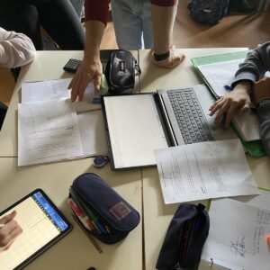 Students sitting around a desk with a laptop, a tablet and some paper sheets and writing utensils.