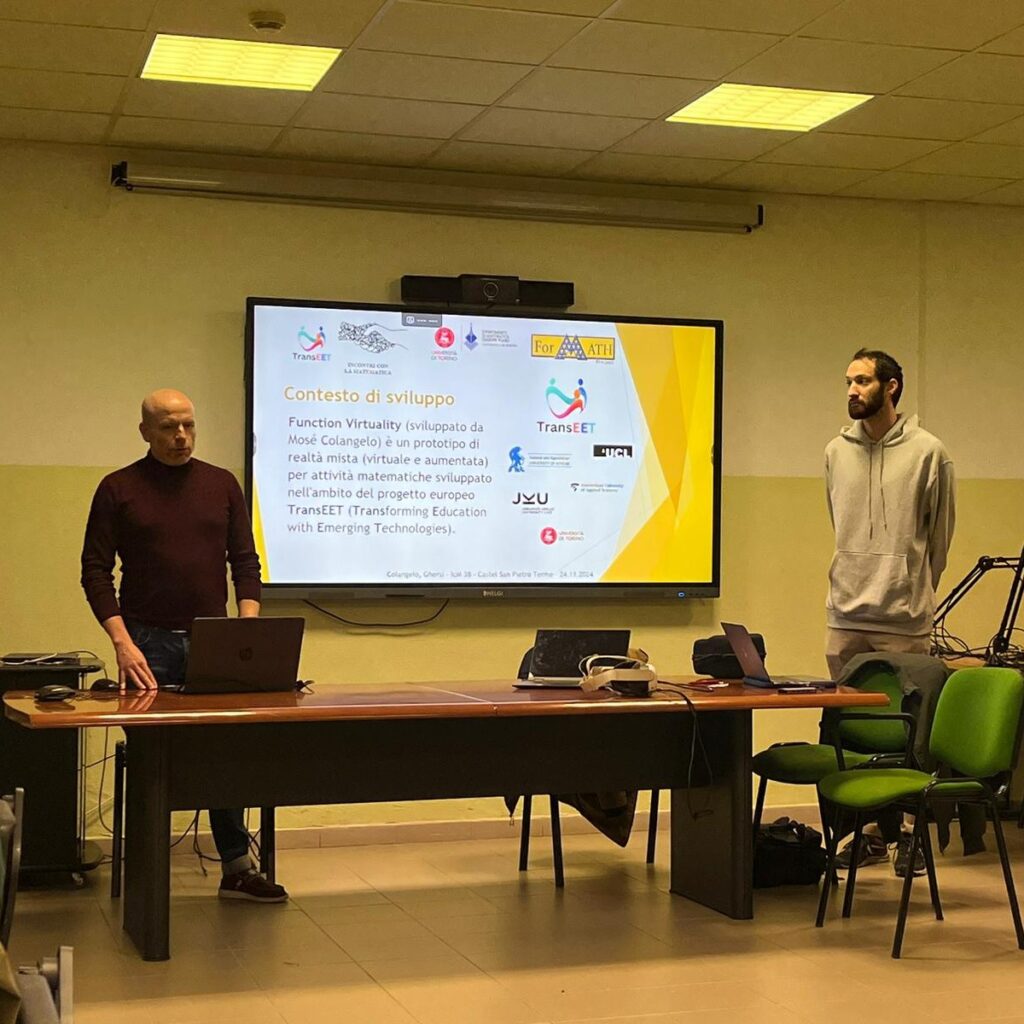 Andrea Ghersi and Mosè Colangelo presenting in a classroom. A big TV screen shows an introduction text of the Function Virtuality application prototype and logos of the involved partner universities and projects.