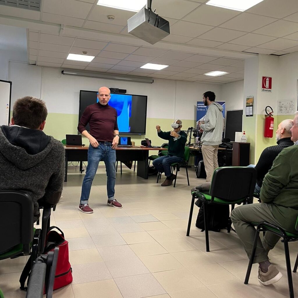 Andrea Ghersi presenting in front of a workshop audience while Mosè Colangelo is supporting a workshop participant who is wearing a virtual reality headset.
