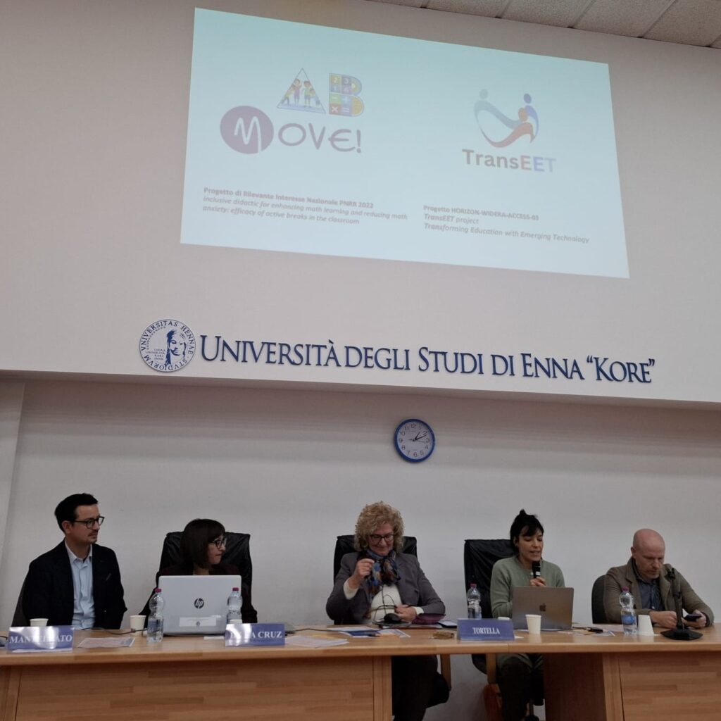 A photo of a panel with five people including Carlotta Soldano and Andrea Ghersi at the Kore University of Enna. In the background the name of the University ‘Università degli studi di Enna “Kore”’ is written in big letters on the wall. Above it a projected image shows the logo of the project move! and the logo of the Transeet project.