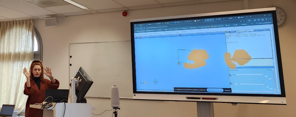 A woman standing at a speaker's podium with a large screen beside her. The screen shows a simple representation of her hands projected over the malt2 software.