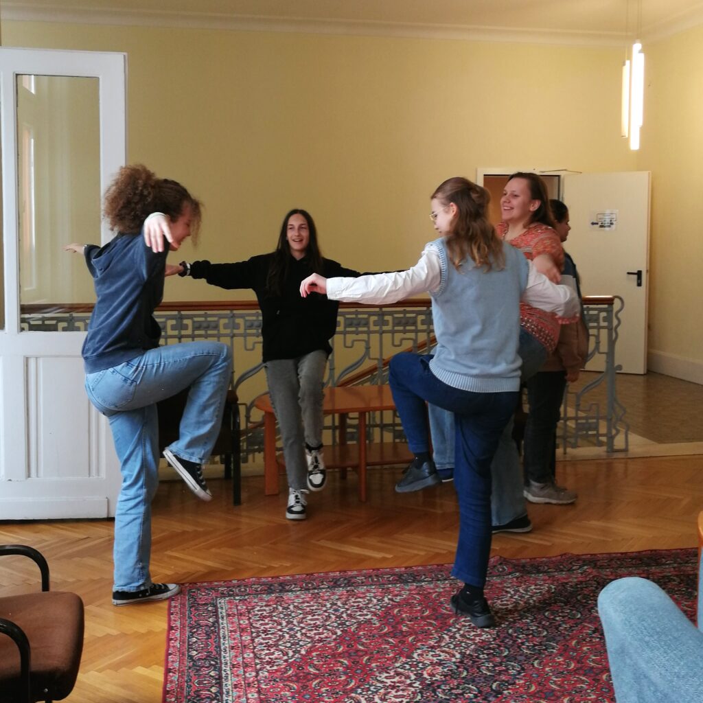 A photo of four girls standing in a circle on one leg with their arms stretched horizontally mirroring each others’ dance movements.