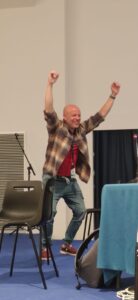 Andrea Ghersi is smiling at the audience with his arms raised above his head and his fists closed, simulating the shape of a parabola.
