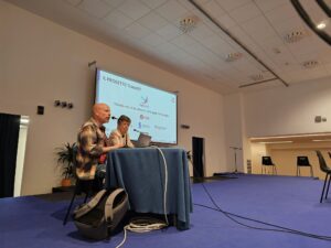 From the left, Andrea Ghersi and Ornella Robutti, are seated at a table and presenting in front of a large screen displaying the title 'Il Progetto TransEET'.