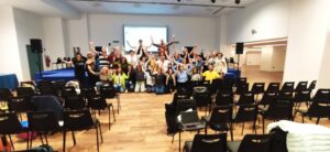 A large group of the workshop participants, with their hands raised simulating a the shape of a parabola, are posing for a photo in a conference room. The group is smiling and standing in front of a projection screen.