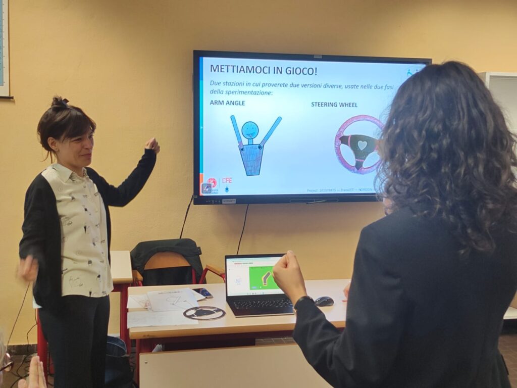 Carlotta Soldano and a workshop participant are gesturing to each other while observing the computer screen. They have their arms raised to try out the angle applet with GeoGebra Plus. The projection screen in the background of the image displays the title ‘LET'S GET INVOLVED!’ and two illustrations side by side entitled ‘ARM ANGLE’ (showing a stylised figure with arms raised) and ‘STEERING WHEEL’ (showing a steering wheel).