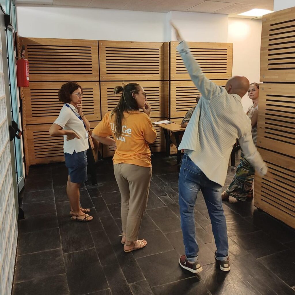 Andrea Ghersi, with his back to the camera, gestures with his arms outstretched to form a straight line, showing a group of participants how to interact with the GeoGebra Plus applet. 
