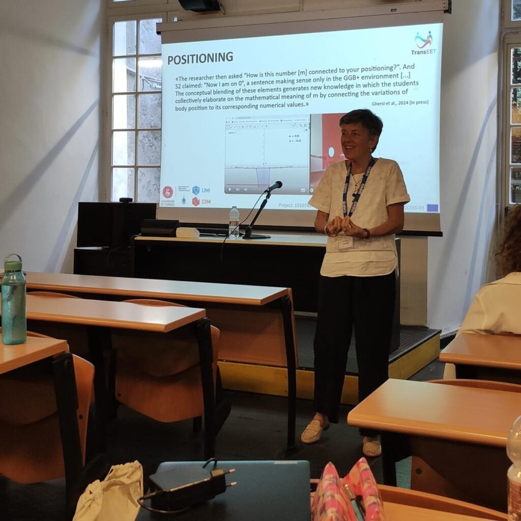 Ornella Robutti is smiling and gesturing as she explains a presentation slide titled 'POSITIONING' to the participants.