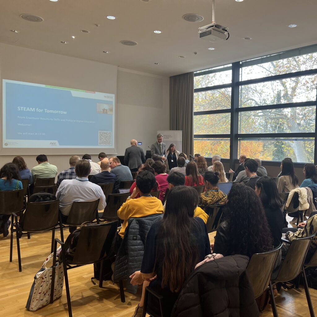 The audience of the STEAM for Tomorrow event sitting in front of projector screen with the name of the event projected on a blue background.