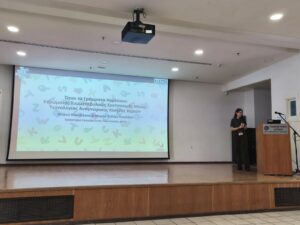 A photo of Myrto Karavakou presenting on a large stage with a slideshow in the background.
