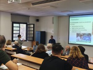 A photo of Manolis Mavrikis and Christina Gkreka discussing in front of a lecture room audience. The projected slide show in the background shows the text “Polarisation. The Enthusiasts: GenAI as educational revolution”